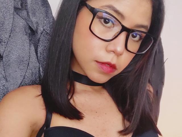 MaddieBlossom with black glasses, red lipstick, black choker, and straight black hair, showing her tits in a black strap bra, looking into the camera.