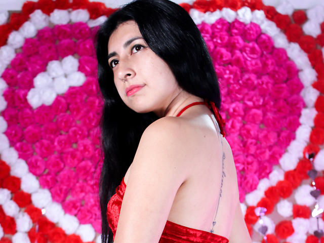 EmmaXWilliams, con cabello negro largo, vestido rojo y un tatuaje visible en la espalda, mirando por encima del hombro, mostrando sus senos, sobre un fondo de un corazón de rosas rosadas y blancas..