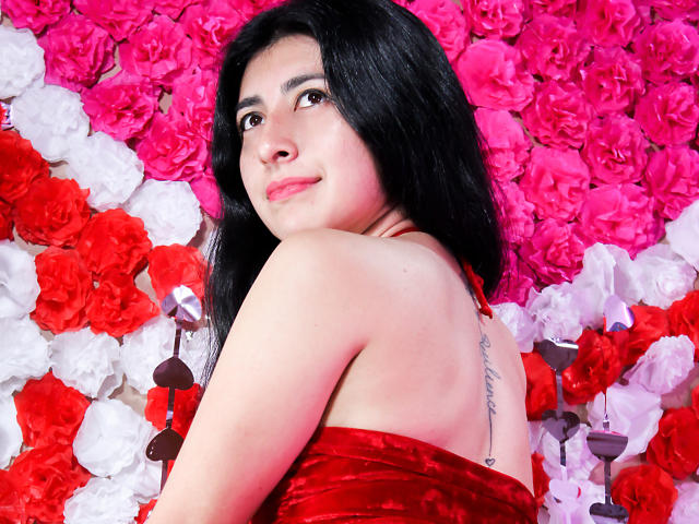 EmmaXWilliams, com cabelo preto, vestindo um vestido de veludo vermelho, sorrindo, olhando por cima do ombro, com uma tatuagem na parte superior das costas, cercada por rosas rosa e vermelhas, segurando óculos de sol..