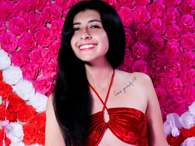EmmaXWilliams, com longos cabelos pretos, sorrindo, vestindo uma blusa vermelha de veludo com alças, que expõe seios pequenos, e com uma tatuagem no lado direito do peito. Rosas rosa e brancas ao fundo..
