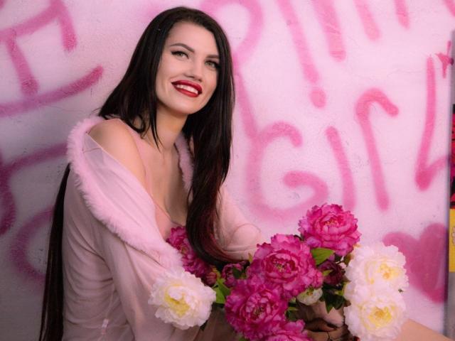 MilaLoona met lang zwart haar, rode lippenstift, een roze bontjas en roze bloemen, lachend naar de camera, terwijl ze een boeket roze en witte rozen vasthoudt..