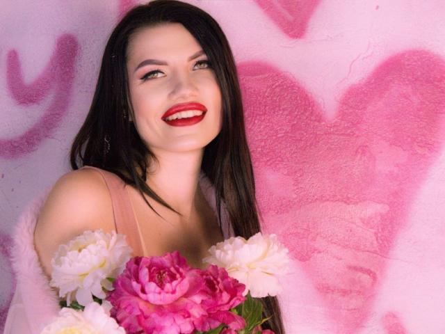 MilaLoona with long black hair, red lipstick, and pink dress, smiling, holding pink and white flowers, tits visible, pink heart graffiti wall, sexy, cute, happy.