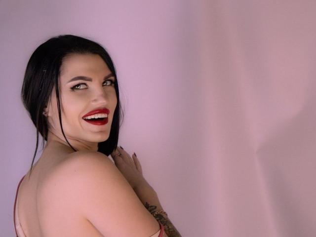 MilaLoona, sem blusa, com cabelo preto, batom vermelho, sorrindo, mostrando tatuagens no braço direito, olhando por cima do ombro, fundo rosa. Seios parcialmente visíveis. Mãos perto do pescoço, expressão brincalhona..