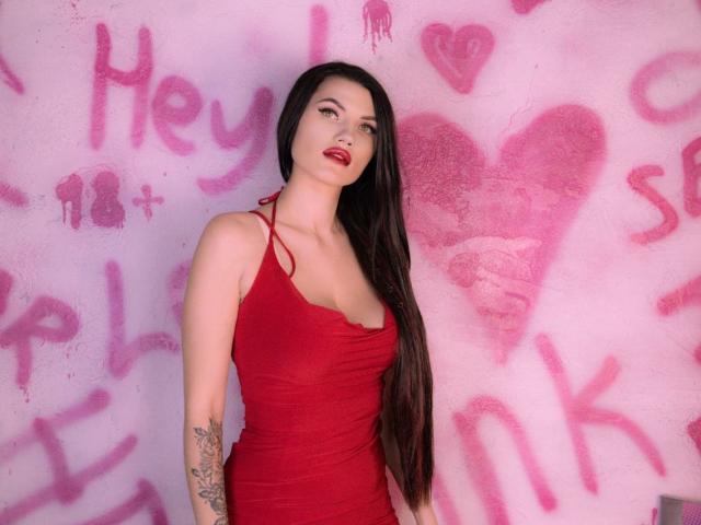 MilaLoona in a red, tight, sleeveless dress, standing with long dark hair, red lipstick, tattoo on left arm, looking at camera. Her tits are visible, she's hot.