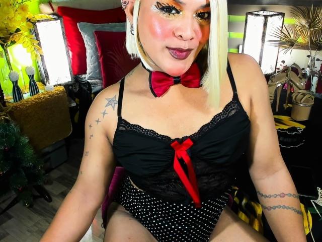 TransGiftedCum with blonde bob, orange eyeshadow, red bowtie, black lace top, polka-dot panties, tattoos, sitting, showing off her tits and ass. Background is blurred.