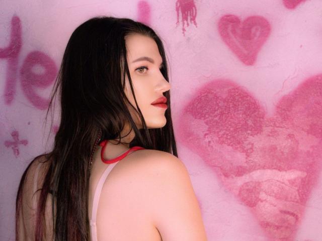 MilaLoona with long black hair, red lipstick, and a pink tank top, showing her back and side profile, looking over her shoulder with a subtle smile.