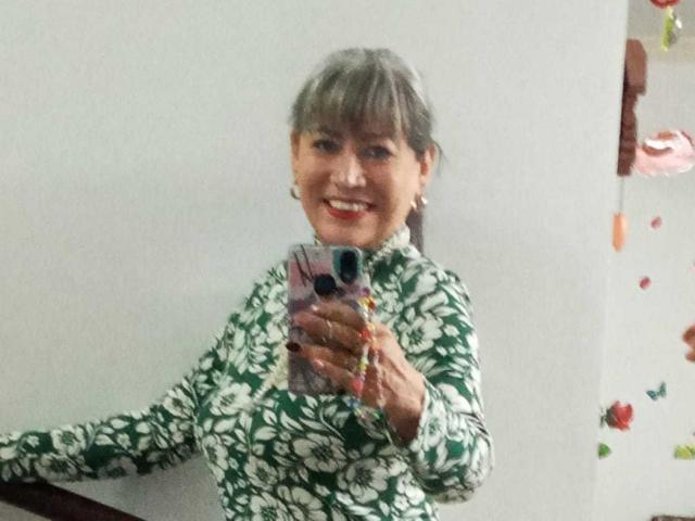 SamanthaLoveLatina, con cabello gris, vestida con un vestido floral verde y blanco, sonriendo, sosteniendo un teléfono rosa, tomando una selfie en el espejo, mostrando sus senos y pintalabios rojo..