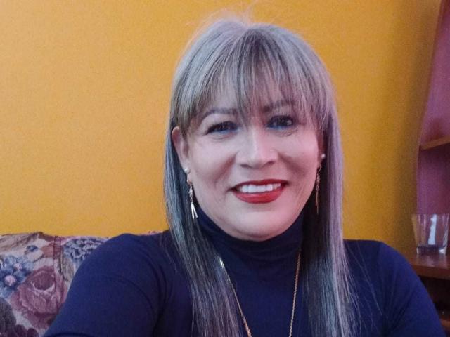 SamanthaLoveLatina, con flequillo rubio, lápiz labial rojo, suéter negro de cuello alto, aretes y collar dorados, sonriendo a la cámara, sentada en un sofá con estampado floral..