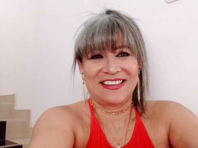 SamanthaLoveLatina hat graue Strähnen, roten Lippenstift und ein Lächeln, trägt ein rotes Top mit Halter, eine goldene Halskette und Ohrringe, und hat ein sichtbares Nasenpiercing..