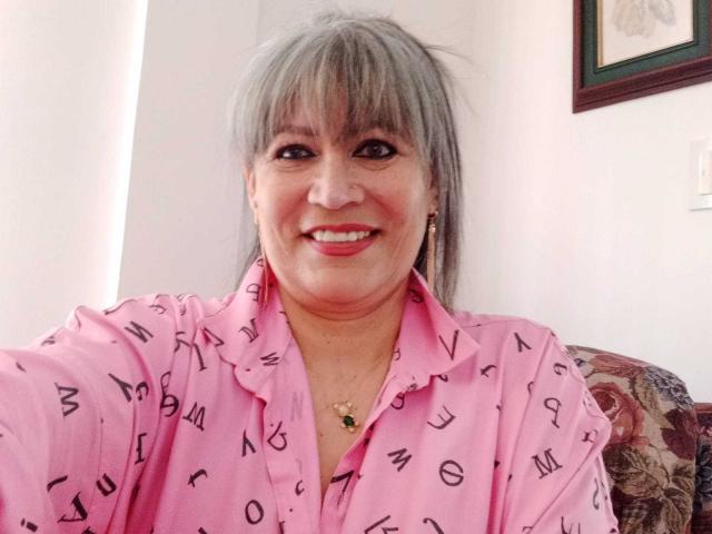 SamanthaLoveLatina with gray hair, smiling, wearing a pink shirt with black letters, small gold necklace with green pendant, red lipstick, and gold earrings, sitting on floral chair.