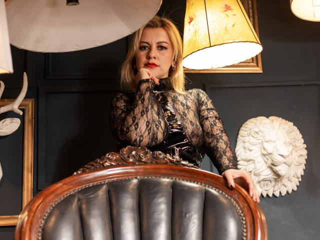 PerlaDivaio with blonde hair, red lipstick, lace top, and black glossy bustier, resting finger on chin, sitting on ornate leather chair, under warm lamps.