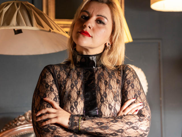 PerlaDivaio with blonde hair, red lipstick, black lace top, and black leather collar, arms crossed, hands with red nails, looking confident, wearing a shiny black choker, in a warm light.