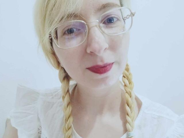 AlexandraSweet has blonde braids, glasses, red lipstick, and a white shirt. Her face is centered, showing a slight smile. Her tits are small, and her skin is fair.