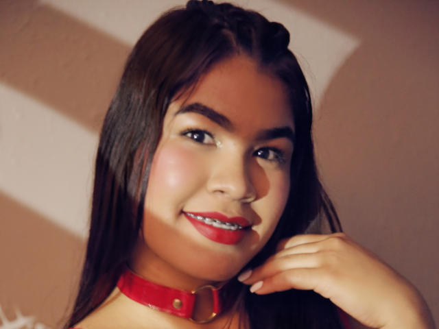 AmyXPlata ha lunghi capelli castani, rossetto rosso e un collare rosso con un anello dorato, sorride con la mano vicino al viso. Si vedono i suoi occhi castani e la sua pelle liscia..