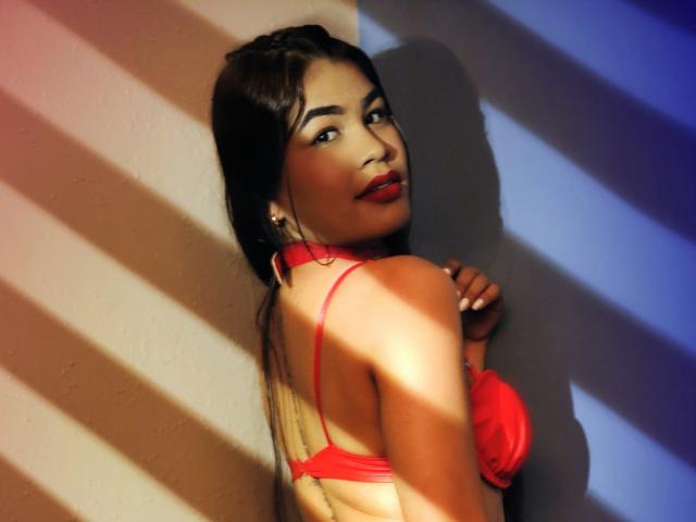 AmyXPlata con reggiseno rosso, che guarda indietro, mani appoggiate al muro, capelli scuri, rossetto rosso, strisce di ombretto, muro color beige chiaro, mostra il seno, sexy, sicura di sé, asiatica, in posa..