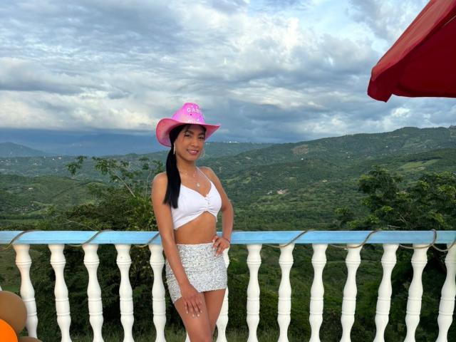 NatashaLewis with black pigtails, pink cowboy hat, white tied-up top, and white floral mini-skirt, standing by a white rail, showing her small tits and smiling.