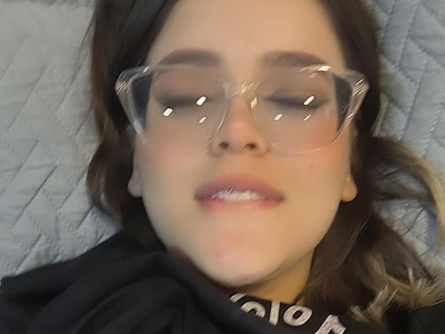 Kim, met haar ogen gesloten en een paar heldere plastic brillen op, heeft een lichte glimlach op haar gezicht en ligt op een lichtgrijze, gewatteerde ondergrond, met haar bruine haar uitgespreid..