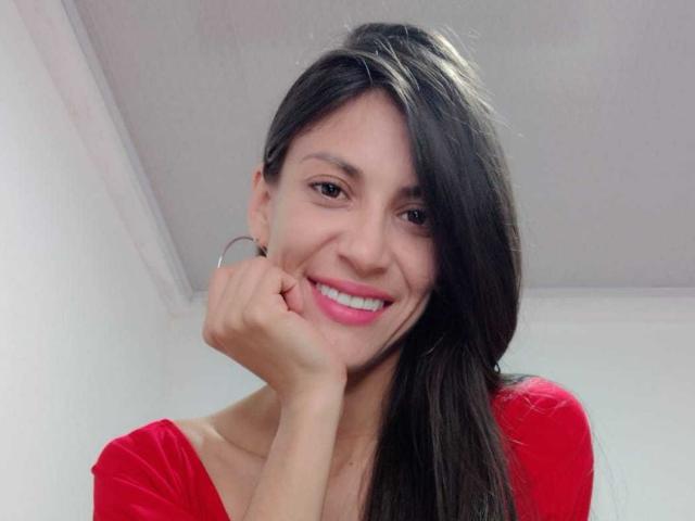 AlejaSexi a de longs cheveux noirs, du rouge à lèvres rose et un haut rouge, elle sourit en posant sa main sur sa joue, elle porte des boucles d'oreilles pendantes, mettant en valeur son joli visage et son sourire séduisant..