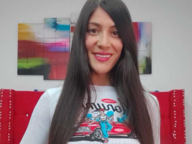 AlejaSexi tiene el cabello largo, lacio y negro, ojos marrones y una sonrisa encantadora con lápiz labial rosa. Lleva una camisa blanca con un estampado de un coche rojo..