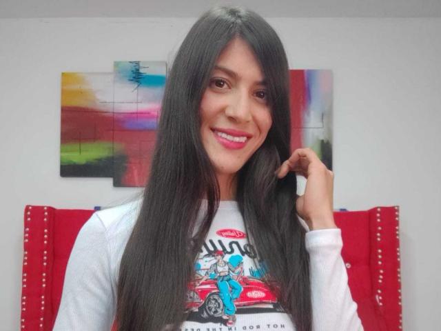 AlejaSexi com cabelo preto longo, sorrindo, vestindo uma camiseta branca com estampa, tocando o cabelo, batom rosa, fundo desfocado vermelho e colorido, tecido vermelho com pontos brancos..