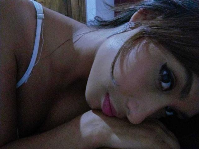VanessaLove with dark hair, wearing a white strap, has her tongue on a man's arm, her eyes looking up, wearing a diamond earring, in a dimly lit room.