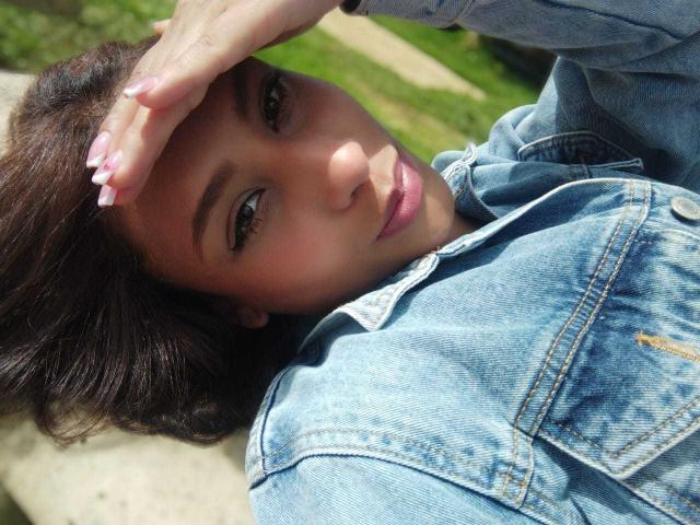 VanessaLove with dark brown hair, green eyes, and pink lips, wearing a blue denim jacket, lying down, hand covering her forehead, sunlight on her face, pink nails.