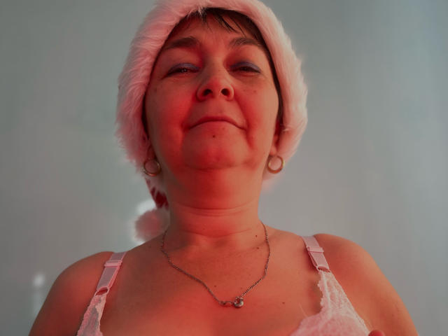 MileyStorm, con un gorro rosa de Papá Noel de textura suave, sacando la lengua, vistiendo una blusa blanca de encaje y un collar de plata, tiene aretes agrandados y un pequeño piercing en la nariz..