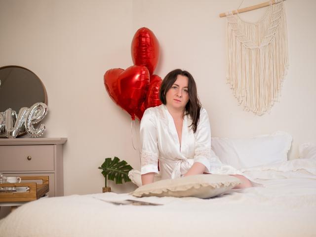 AbbyTaylorr sits on a bed in a white silk robe, showing her small tits, with red heart balloons behind her and a macramé wall hanging.