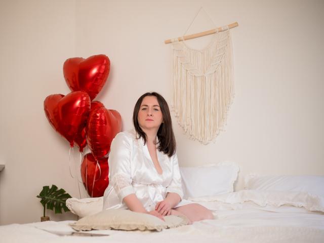 AbbyTaylorr, sitting on a bed, wearing a white silk robe, with red heart-shaped balloons and a macramé wall hanging, has small tits and a cute face, dark brown hair.
