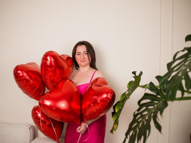 AbbyTaylorr with shiny red heart balloons, wearing a pink dress, holding them in front of her, standing next to a green plant, smiling slightly.