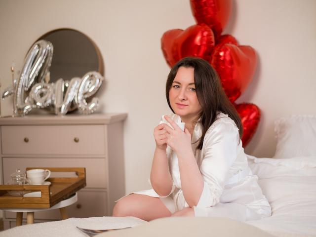 AbbyTaylorr with brown hair, white robe, sitting on bed, holding hands, cute smile, red heart balloons behind her, white dresser with "love" letters on left.