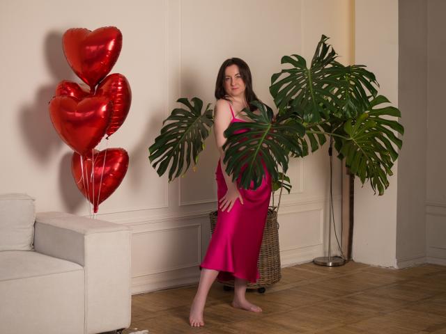 AbbyTaylorr in a pink satin dress, standing barefoot, holding a green monstera plant, with red heart balloons to the left, smiling at the camera.