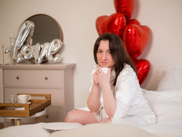 AbbyTaylorr with long brown hair, white robe, holding white mug, sitting on bed, red heart balloons, wooden tray with cup, white dresser with mirror, soft smile, cozy room.