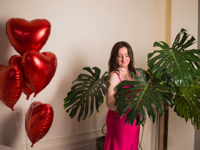 AbbyTaylorr stands in a pink dress, holding a large green monstera leaf, with red heart-shaped balloons to the left, and a green plant to the right.