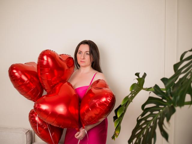 AbbyTaylorr with red shiny heart balloons, wearing a pink dress, holding them against her chest, standing next to a green plant, looking at the camera.