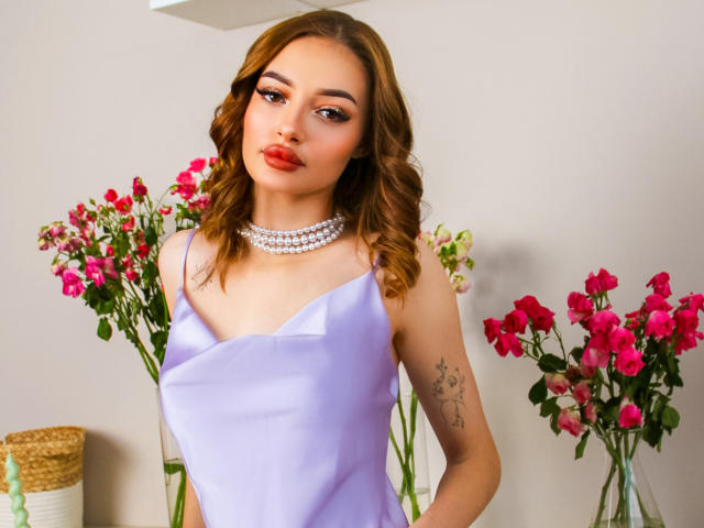Kylie Beaty mit welligem, braunem Haar, rotem Lippenstift und einem lila Seidenkleid, die eine Perlenkette trägt und ein Tattoo auf ihrem rechten Arm zeigt, mit rosa Rosen im Hintergrund..