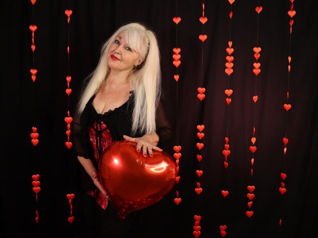 KamaliaSim with long blonde hair, black dress, and red ribbon, holding shiny red heart-shaped balloon, surrounded by hanging red hearts, smiling with red lipstick.