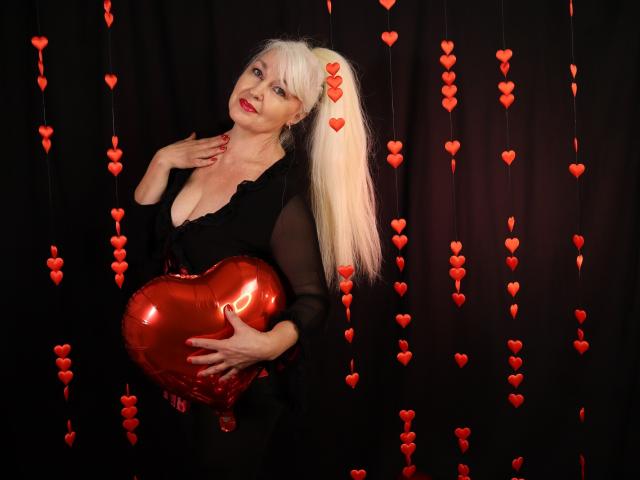 KamaliaSim mit blondem Pferdeschwanz, schwarzem, durchsichtigen Kleid, hält ein glänzendes rotes Herz, rote Herzdekorationen, schwarzer Hintergrund, lächelnd, Hand auf der Brust, roter Lippenstift, helle Haut, süß..