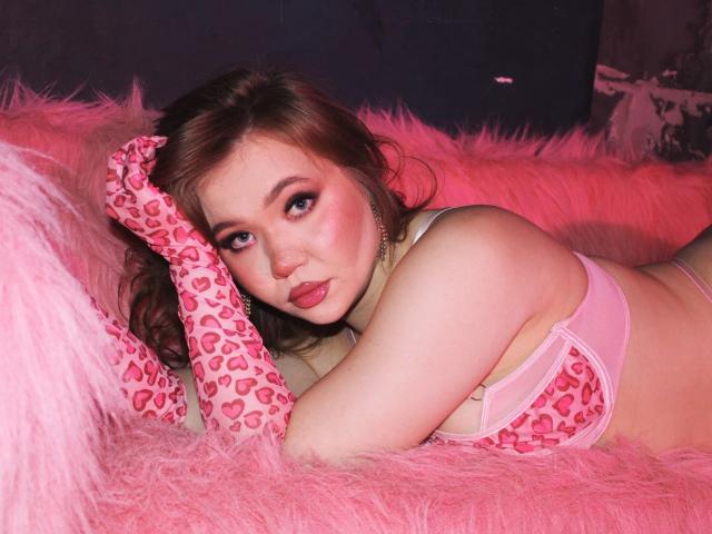 StacyYellig lying on pink furry blanket, wearing leopard-print bra and pink heart-patterned gloves, looking at camera with pink lipstick, showing her ass, and red eyes.