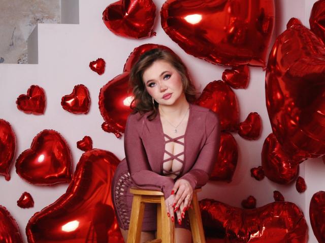 StacyYellig with wavy brown hair, pink lipstick, and red eyeshadow, wearing a pink lace-up dress, leaning on a wooden stool, surrounded by shiny red heart balloons.