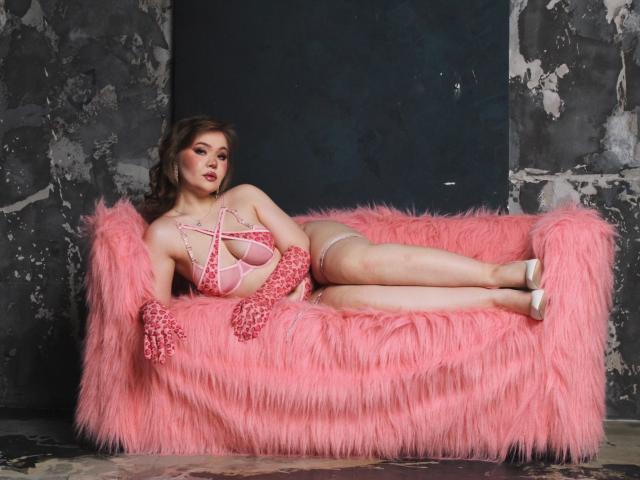 StacyYellig lounges on a pink furry couch, wearing a pink strappy lingerie set with beads, showing her big tits and curvy ass, with pink gloves and white heels.