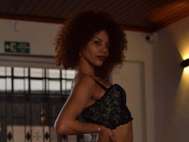 ChanelDupont with curly brown hair, dark skin, wearing black lace bra, standing in dimly lit room, looking over shoulder with subtle smile, small round earrings, wooden ceiling.
