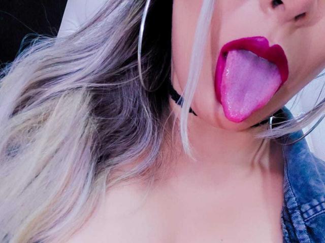 CamilaBardot's tongue is out, wet and pink, with bright red lipstick. She's wearing a black choker and has long, blonde-streaked hair. Her boobs are slightly visible.