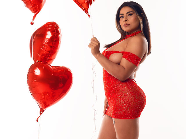 SusanClaire con un vestido rojo, brillante y con aberturas, sosteniendo globos con forma de corazón rojo, mostrando sus curvas y su busto grande, con cabello largo y castaño, sobre un fondo blanco..