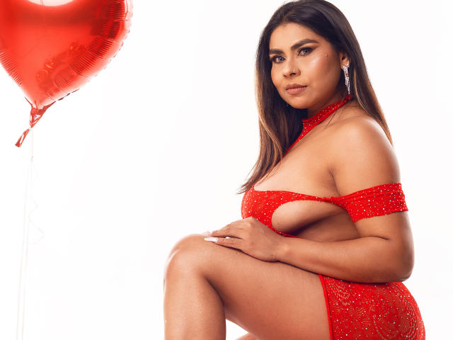 SusanClaire indossa un abito rosso, scintillante, con tagli che mostrano il seno e il lato dei glutei, con lunghi capelli castani, orecchini e un palloncino rosso a sinistra..