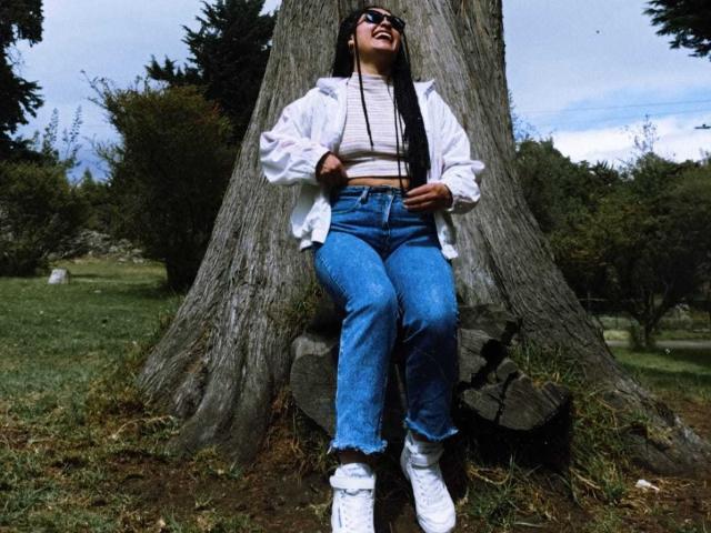 Tokiozz seduto su un ceppo d'albero, indossa una giacca bianca, una maglietta a righe, jeans blu a vita alta, scarpe da ginnastica bianche e occhiali da sole, con lunghi capelli neri, e sta ridendo..