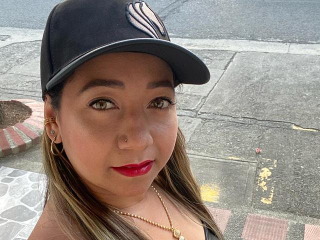 KlhoeHouston has brown eyes, red lipstick, and a nose ring. She wears a black cap with a white logo, gold hoop earrings, and a gold necklace.