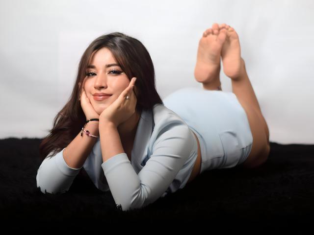 CrystalJoness is lying on a black surface, wearing a light blue dress, with her ass up and feet in the air, hands under her chin, smiling.