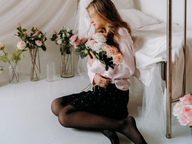 TinaRedly, with long blonde hair, sits on white floor, holding pink roses, wearing pink shirt, black polka-dot skirt, black tights, next to white bed with metal frame.