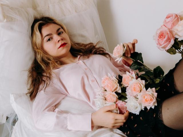 TinaRedly está deitada em uma cama, vestindo uma camisa rosa de botões, uma saia preta e meias, segurando rosas rosa e brancas. Ela tem batom vermelho e cabelo loiro ondulado..
